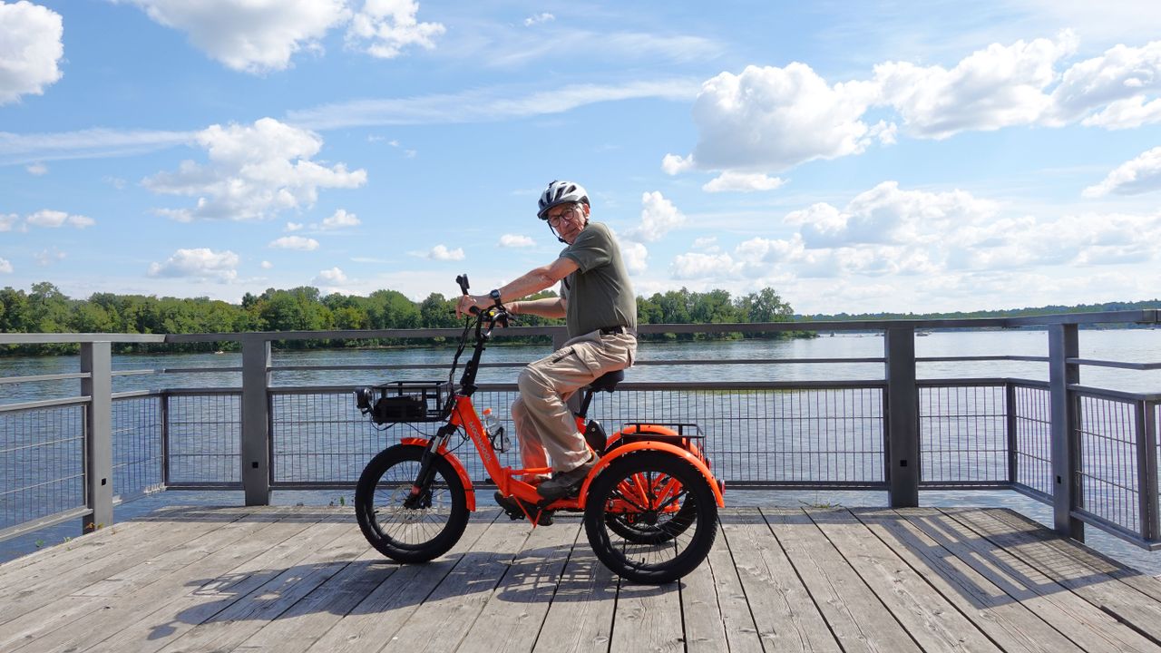 Elderly man enjoying a ride on an electric tricycle by the waterfront on a sunny day.